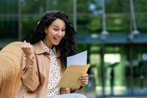 Joyful woman sitting outside, fist pumping while reading excellent news in a letter, expressing excitement and happiness. photo