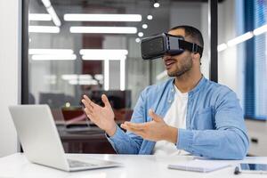 un joven musulmán hombre es sentado en un moderno oficina a un escritorio vistiendo un virtual máscara y teniendo un charla en línea. gesticulando con manos. foto