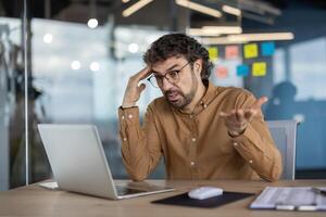 A bewildered businessman in a modern office expressing confusion and frustration while working on a laptop. photo