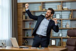 Energetic mature businessman in a smart suit celebrates victory with a fist pump in a well-appointed home office setting. photo