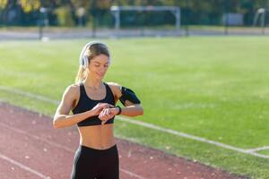 A focused female runner with headphones tunes her workout, checking her smartwatch on a sunny track field. photo