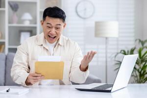 Happy and successful asian received mail notification envelope with good news, man sitting at table at home, working in home office in living room, joyfully celebrating winning news. photo