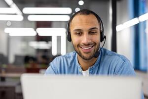 A cheerful male customer service representative wearing a headset with a microphone, working on a computer in a contemporary office setup. photo