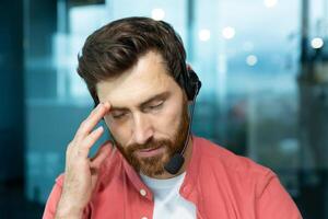 cámara web vista, remoto llamar, triste hombre con auriculares sentado tarde a lugar de trabajo, sobrecargado de trabajo y sobrecargado de trabajo cliente apoyo obrero. foto