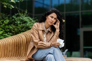 Frustrated caucasian woman sitting on outdoors bench and feeling pain. Attractive female with curly hair holding head with one hand and napkin in other. Concept of ache and diseases. photo