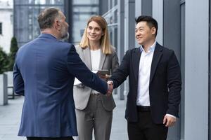 Meeting of three successful business people, diverse dream team man and woman outside office building, greeting and shaking hands, experienced professionals specialists in business suits talking photo