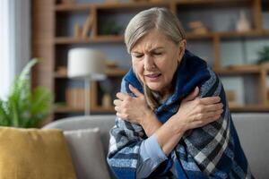 An elderly woman experiencing chills, sitting on the sofa wrapped in a cozy blanket, looking visibly cold and uncomfortable. photo