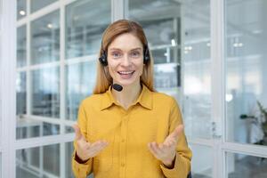 Mature adult business woman with blond hair working inside light office, customer service tech worker smiling and talking to customer consulting, using headset and laptop for remote call. photo