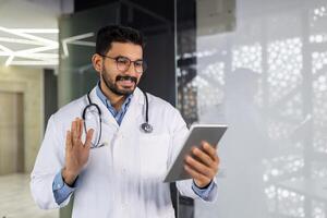 un joven médico sonrisas con un tableta computadora en su manos, negociaciones remotamente con un paciente, consulta utilizando un juego, olas y saluda, soportes dentro el clínica por el ventana. foto