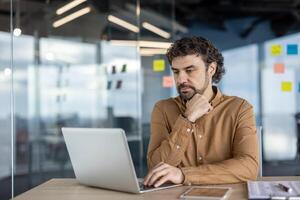 A serious Hispanic businessman deep in thought while working at his laptop in a modern office setting. photo