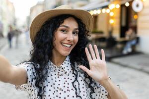 Young beautiful Latin American woman walking in evening city, woman looking into smartphone camera, using call app, talking with friends smiling. photo