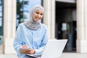 un joven musulmán mujer enfocado en estudiando afuera, tomando notas junto a su ordenador portátil a un Universidad instalaciones. foto