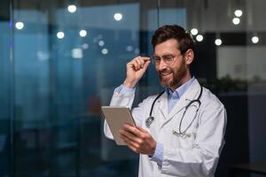 A young male doctor, intern, intern is standing in an office in a hospital in a white coat and with a stethoscope. He works on a tablet, holds his glasses in his hand, and smiles. photo