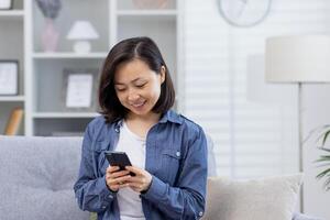 un joven hermosa asiático se sienta solo a hogar en el sofá, el mujer usos un solicitud en el teléfono, sostiene un teléfono inteligente en su manos, sonrisas felizmente, usos el solicitud, navega en línea paginas foto