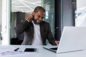 Sick tired man at workplace, businessman has severe neck pain, long sitting work, overworked african american man working long time on project inside office with laptop. photo