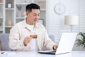 Joyful successful asian businessman working from bright home office, man using bank credit card and laptop for online shopping in online store, and booking services remotely. photo