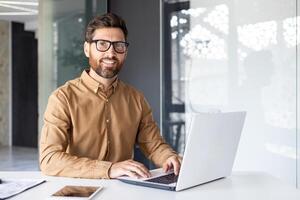 retrato de exitoso hombre a lugar de trabajo dentro oficina, experimentado sonriente empresario en camisa sonriente y mirando a cámara, programador desarrollando nuevo software con ordenador portátil. foto