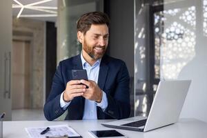 joven sonriente empresario trabajando en el oficina, sentado a el escritorio en un traje, mirando a el ordenador portátil pantalla y participación un móvil teléfono. foto