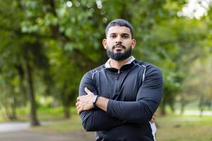 A confident male stands with arms crossed against a lush park backdrop, exuding strength, and determination outdoors. photo