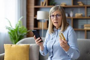 A senior woman is indoors holding a credit card and smartphone, potentially experiencing fraud. photo