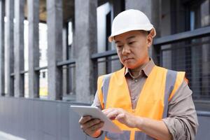 Asian young man, construction worker, foreman, company owner stands outside in hard hat and vest and concentrates on tablet. photo