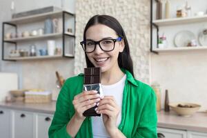 Young beautiful brunette woman in glasses and green shirt at home in kitchen, eating chocolate photo