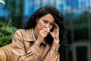 A distressed businesswoman in professional attire is sitting outside, wiping away tears with a tissue, a look of sadness and worry on her face amidst an urban backdrop. photo