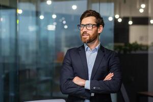 Portrait of successful mature investor, senior businessman inside office looking towards window smiling with crossed arms, man in glasses and business suit. photo