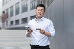 Portrait of a young asian man in a white shirt standing on the street near a modern office center, holding a phone and a credit card, looking at the camera. photo