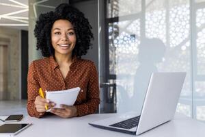 un alegre joven africano americano mujer con Rizado pelo con confianza trabajos a su ordenador portátil en un brillante, moderno oficina ajuste. foto