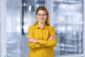 Portrait of successful mature business woman inside office, female worker in yellow shirt smiling and looking at camera with crossed arms. photo