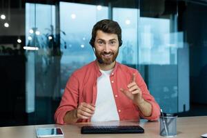 call, joyful smiling man with headset phone working in evening time inside office, web camera view, online meeting with colleagues partners remotely. photo