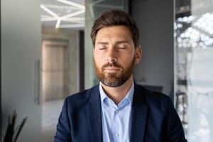 de cerca retrato de un joven hombre trabajando en el oficina, sentado pensativamente en un traje con cerrado ojos y descansando foto