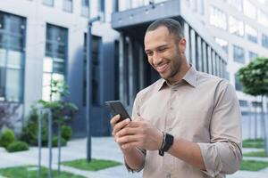 Young african american businessman using smartphone, smiling and happy typing message online, man outside office building typing message to friends photo