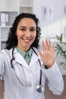 vertical disparo, hembra médico sonriente alegremente ondulación a teléfono cámara, hablando a pacientes remotamente utilizando teléfono inteligente llamada aplicación, trabajando dentro clínica oficina. foto