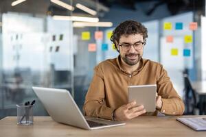 Casually dressed businessman working with a digital tablet and laptop at a wooden desk in a contemporary office setting. Productive and relaxed atmosphere. photo