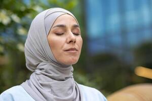 un sereno retrato de un musulmán mujer meditando con cerrado ojos, vistiendo un hiyab, en un verde urbano parque, exudando un sentido de paz y contentamiento. foto