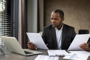 Sceptic formal male in business suit looking attentively on sheets of paper in both hands while sitting by desktop. Focused accountant comparing data on two documents from different departments. photo