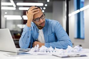 Exhausted Indian businessman feeling stressed with crumpled papers, laptop in office setting. Overworked office worker concept. photo