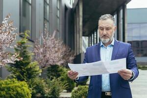 Real estate agent. Handsome senior serious man in suit and beard standing on street near houses, office center, holding documents in hands, plan of apartments. photo