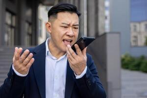 Close-up photo of angry young asian man businessman in suit standing outside and emotionally talking on the phone through loudspeaker, unhappy recording voice message.