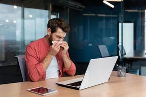 enfermo hombre a trabajo tiene gripe y frío, empresario estornudos y toses a lugar de trabajo trabajando dentro oficina a escritorio utilizando ordenador portátil a trabajar. foto