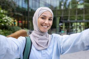 un alegre musulmán mujer en un hijab capturas un autofoto, radiante felicidad, con un oficina edificio fondo. foto