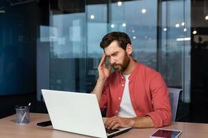 Sick man at workplace, mature businessman has severe headache, boss in shirt works inside office with laptop. photo