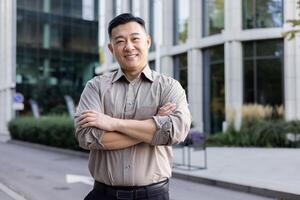 Portrait of a young Asian man standing near an office center, with his arms crossed on his shoulders and looking at the camera, with a smile. photo