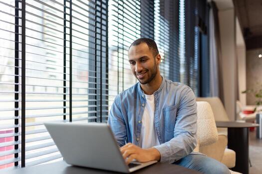 sonriente indio hombre a hogar utilizando un ordenador portátil en un iluminado por el sol vivo habitación, representando remoto trabajo y comodidad. foto
