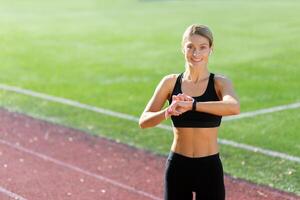 un joven mujer atleta en pie a el estadio vistiendo auriculares y sonriente a el cámara, comprobación el hora en un aptitud pulsera, mirando a el hora de correr, clases foto