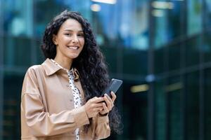 Portrait of a successful and satisfied Latin American business woman, female boss smiling and looking at the camera, holding a smartphone, using an online application, dialing a message and a call. photo