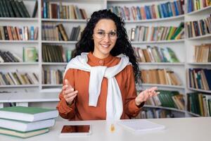 A young woman actively engaging with someone unseen, using expressive hand gestures, sitting in front of a bookshelf with books. photo