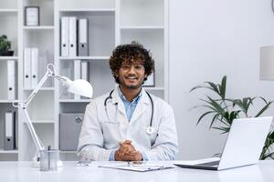 Portrait of young Indian doctor, man in white medical coat smiling and looking at camera, doctor sitting at table inside medical office of clinic, working with laptop. photo